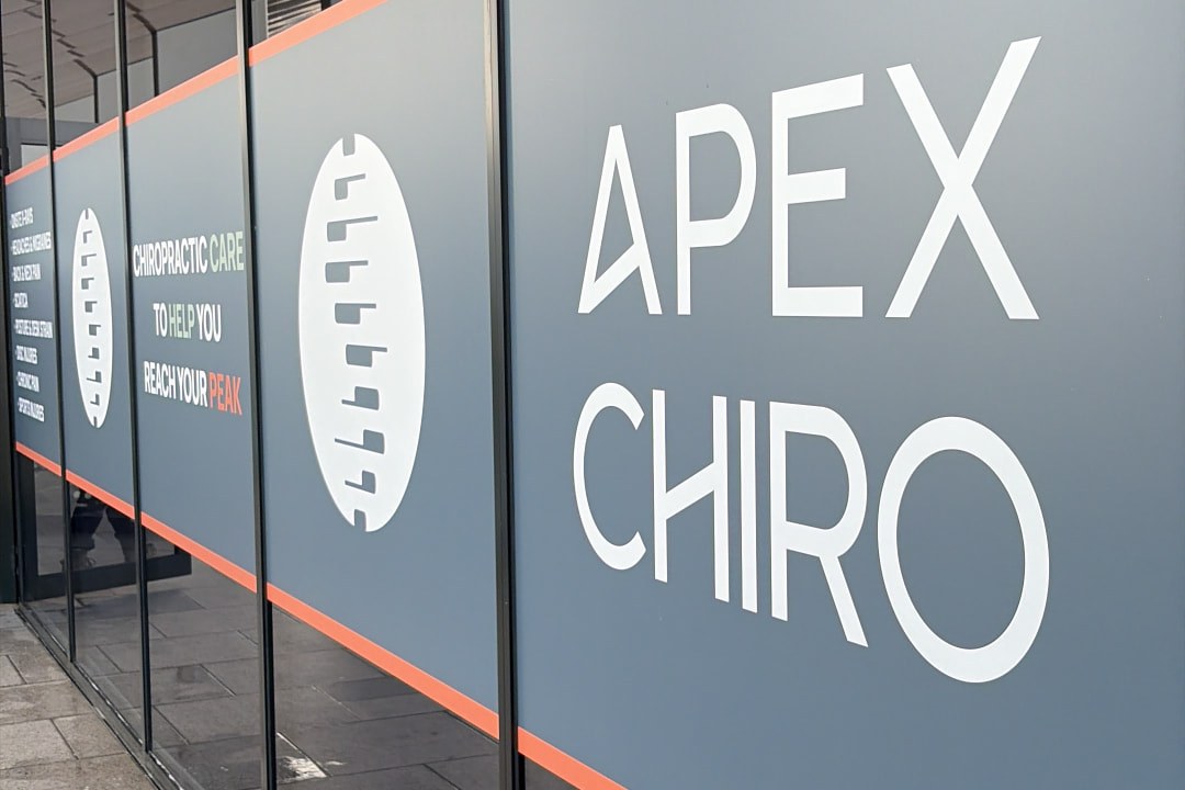 APEX. Chiro Gregory Hills clinic windows facing the walkway at The Hub on Lasso Road, showing the spine logo and the tagline chiropractic care to help you reach your peak